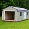 A 12x32 Frontier Garage Shed, featuring a beige and brown exterior with a metal roof, is situated on a grassy field. It includes a garage door at one end and a standard door on the side. The natural green backdrop of trees enhances the setting.