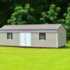 A 12x32 Frontier Garage Shed with a beige exterior, gray roof, and white double doors is positioned on a vibrant green lawn. Each side of the doors features two small windows with black frames. The serene outdoor setting is completed by lush trees forming a backdrop.