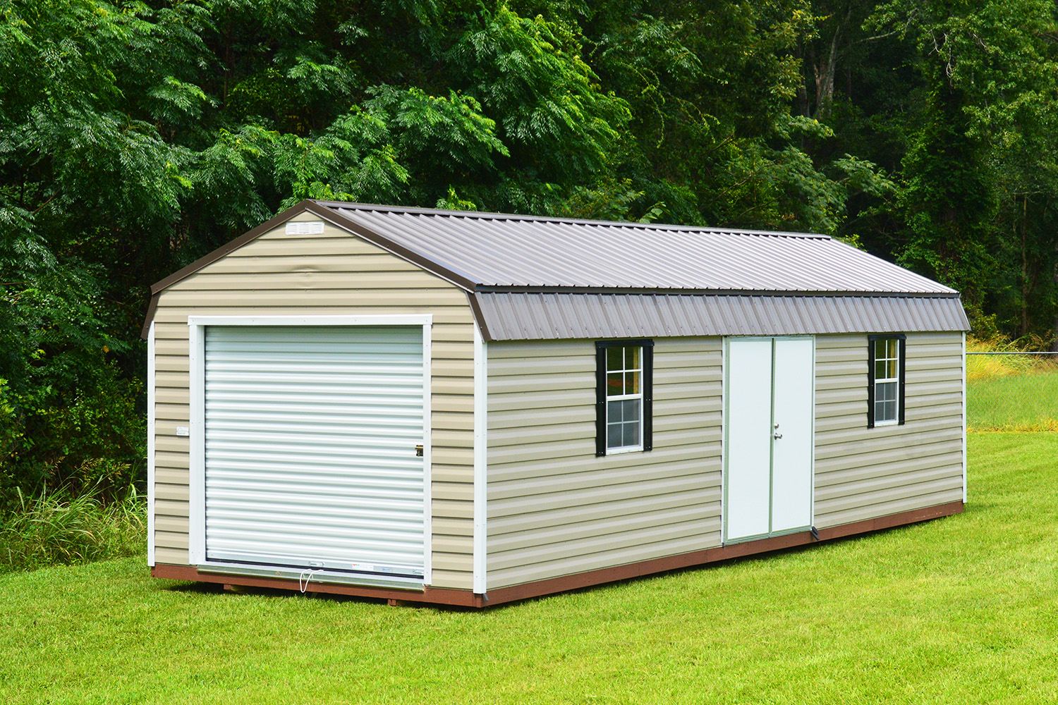 A 12x32 Frontier Garage Shed featuring a light brown rectangular design with a dark metal roof and a white garage door stands on a grassy field. Two small windows and a side entrance door are visible, with tall green trees forming the background.