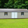 The 12x32 Frontier Garage Shed, featuring a compact beige design with a durable metal roof and two windows, is situated on a grassy lawn with lush green trees in the background.