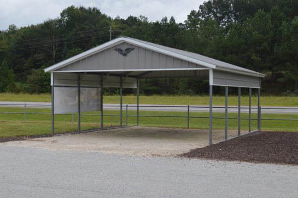 A compact, open 18x20x7 Vertical Carport with a peaked roof and an eagle emblem at the top stands on a gravel surface beside a road, surrounded by grass and trees under a cloudy sky.