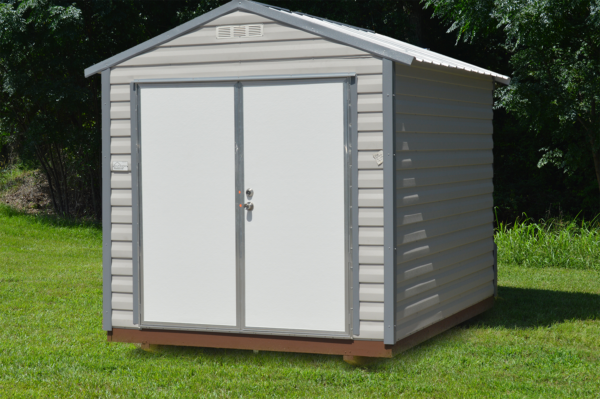 The 8x10 Ridgeline Shed, featuring a compact beige metal construction with a peaked roof and double doors, is positioned on a green lawn amidst surrounding trees.