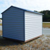 An 8x10 Ridgeline Shed, featuring a small footprint with metal cladding and a blue roof, is situated on a gravel area. It's surrounded by grass and pavement, with a wooded area in the backdrop under a partly cloudy sky.