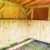 The interior of the Pasture Shelter & Tack Room features exposed beams and unpainted wooden panels. Sunlight filters through the openings, illuminating the grass floor. A small hatch and a mounted paper notice are visible on one wall.