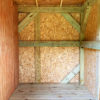 The interior of the Pasture Shelter & Tack Room features light-colored wood paneling and cross-bracing on the walls. Both the flooring and walls are constructed from wood, with a partially visible door on the left side. The space is illuminated by natural light.