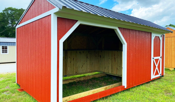 The Pasture Shelter & Tack Room features a red wooden design with a black metal roof and white trim. It includes an open front entrance and a side door adorned with a crossbuck pattern. The shelter is situated on green grass, surrounded by similar structures against a backdrop of partly cloudy skies.