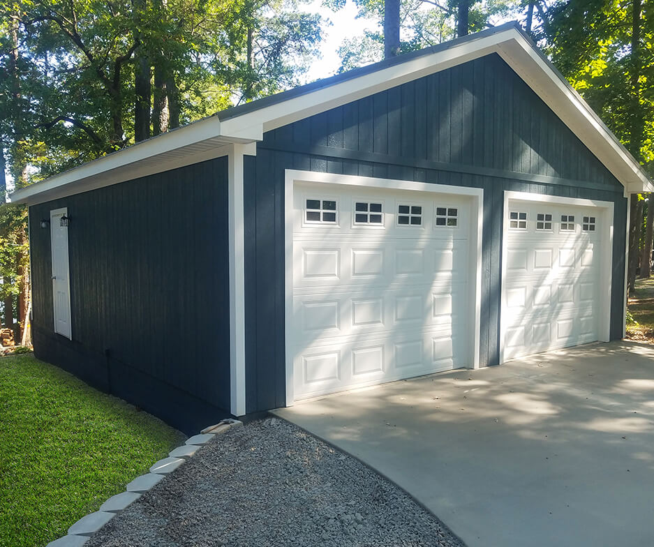 A dark blue detached garage with two white doors stands on a paved driveway. The garage is surrounded by greenery and trees, with sunlight filtering through the leaves, casting soft shadows on the ground.