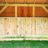 A Pasture Shelter with an open front and a slanted roof stands on a grassy area. The structure features natural wood paneling and beams, with an opening at the bottom front. Grass and wildflowers surround its base.