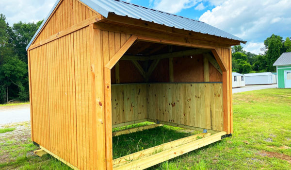 A Pasture Shelter featuring a slanted metal roof and framed with exposed wooden beams is situated on a grassy area. Trees and other small structures are visible in the background under a partly cloudy sky.