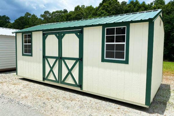 The 10x20 Ridgeline Shed, featuring a cream exterior with green trim and a green metal roof, is positioned on a gravel path. It includes double doors adorned with a window pattern and two large side windows. The scene is complemented by trees and a cloudy sky in the background.