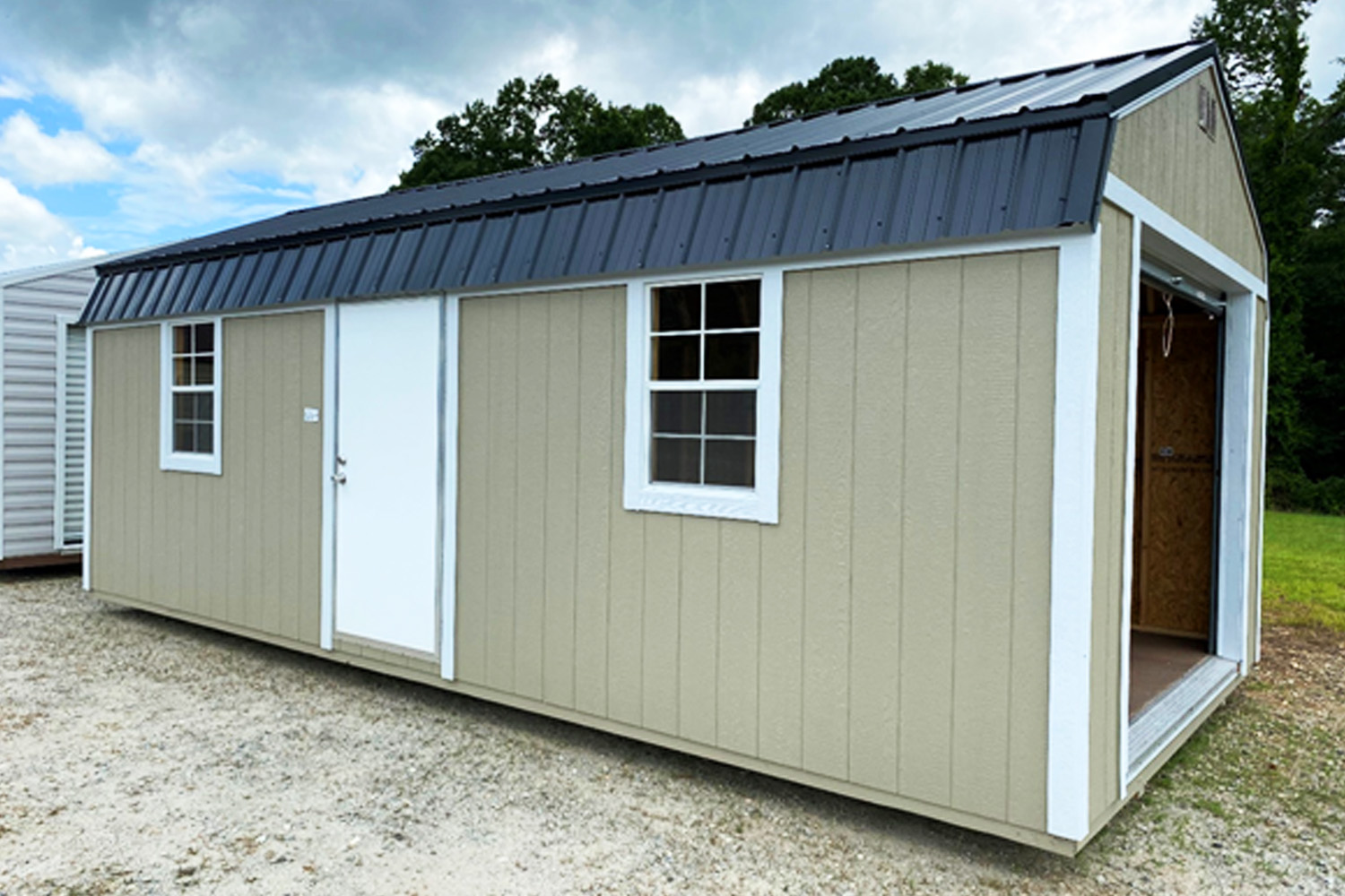 A 12x24 Frontier Garage Shed, featuring beige and white walls, a black metal roof, and a gravel foundation, is equipped with a white door, two windows, and an open side entrance. Trees stand under the backdrop of a cloudy sky.