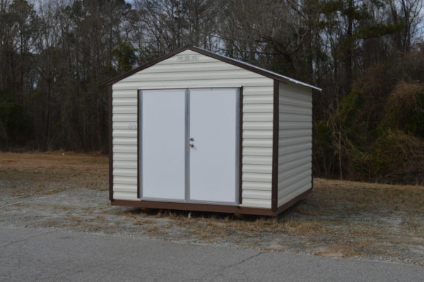 A 10x10 Ridgeline Shed, featuring a white and brown metal exterior, is situated on a grassy patch adjacent to a road, with trees and a wooded area set against a cloudy sky in the background.