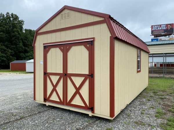 A 10x20 Frontier Shed featuring a tan exterior is equipped with a striking red metal roof and red-trimmed double doors, situated on a gravel lot. It includes a small window on the side, while a billboard can be seen in the background.