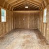 Interior of an unfinished wooden shed with plywood walls and ceiling. Two windows on either side allow natural light to illuminate the spacious room. The floor shows wood grain patterns, enhancing the rustic appearance. A note is pinned to the far wall.