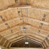 View of an unfinished wooden shed interior with a vaulted ceiling. The structure shows exposed wooden beams and plywood panels. A small window is visible at the back wall, and there is a single light fixture hanging from the ceiling.