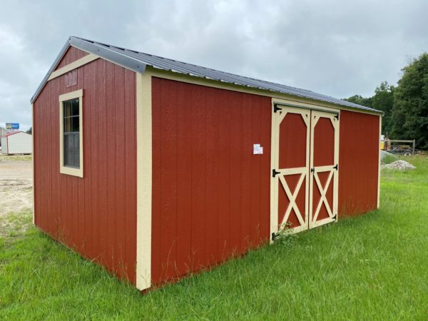 A 12x20 Ridgeline Shed, featuring a red and beige wooden exterior with a metal roof, stands on a grassy area. It includes one window on the side and has double doors at the front with a diagonal cross design. The sky is overcast above it.