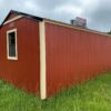 A small red shed with beige trim sits in a grassy field under a cloudy sky. A sign on the roof reads CALL ME ALUMINUM. Trees and a building are visible in the background.