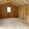 An empty, unfinished wooden interior of a small shed or cabin with a sloped metal roof. The space has plywood walls and flooring, and a single window on the far wall allows natural light to enter.