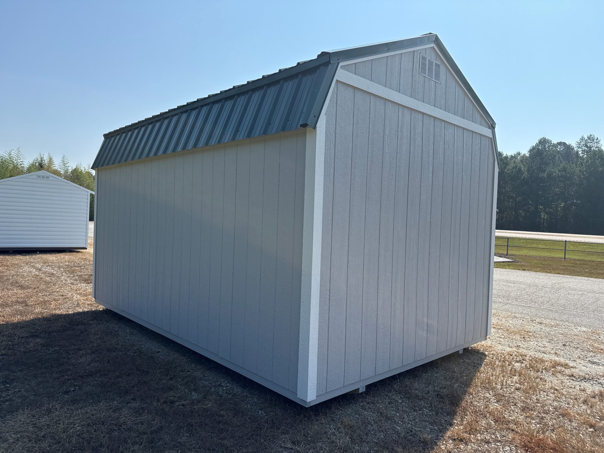 A 10x20 light gray Frontier Shed with a dark roof sits on dry grass by a road, another shed and trees visible under blue sky.
