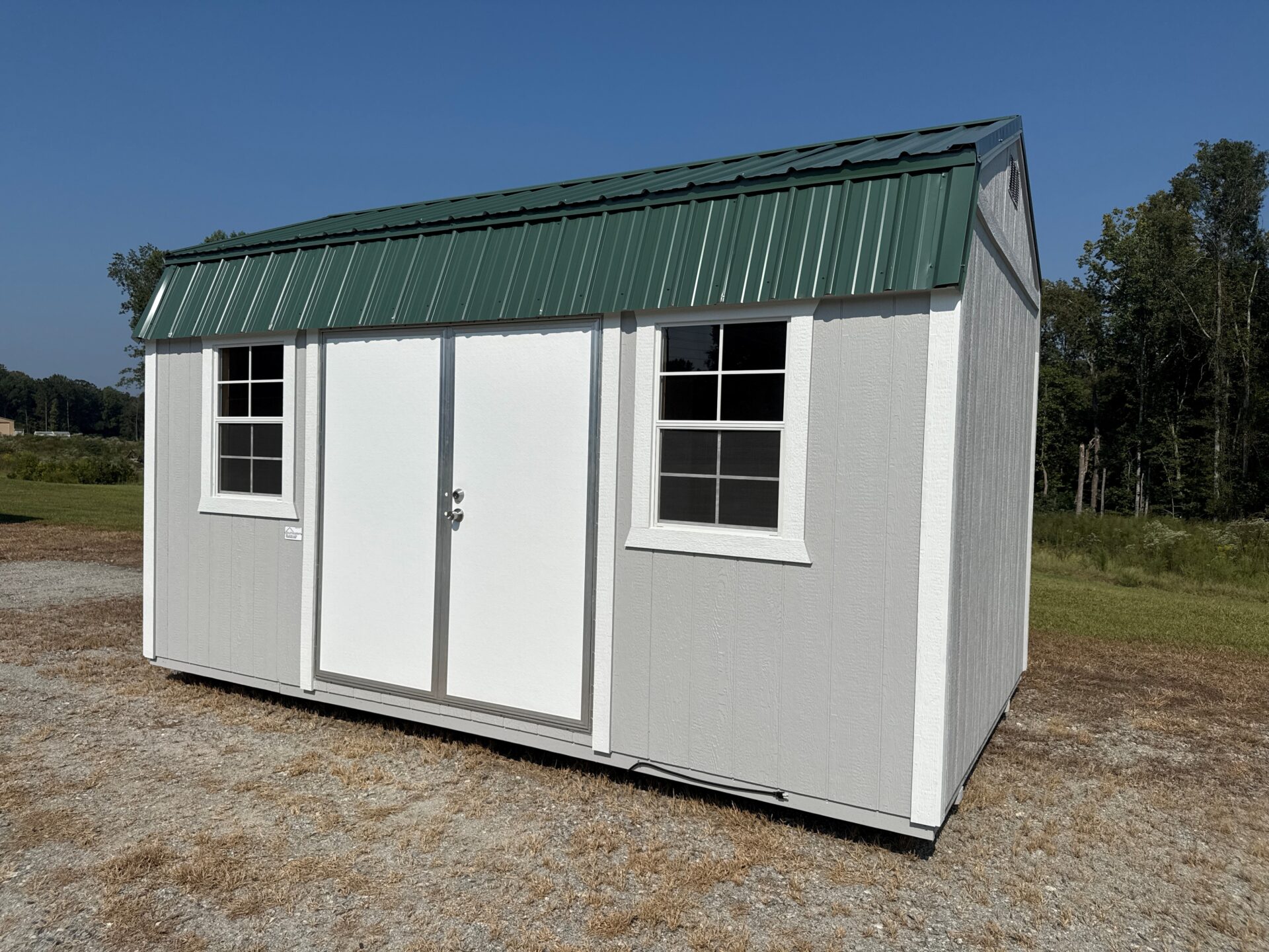 The 10x20 Frontier Shed has a white exterior, green metal roof, double doors, and two windows. It sits on gravel by grass and trees.