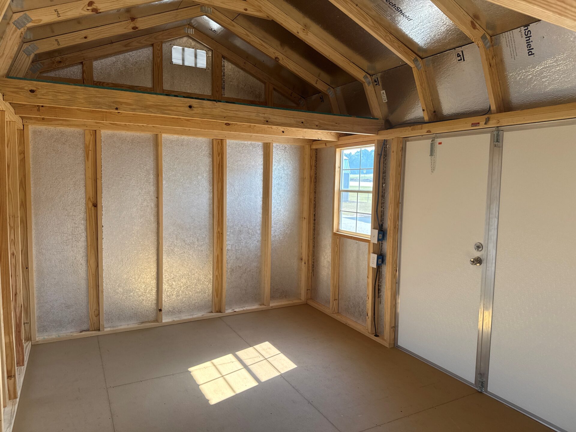 10x20 Frontier Shed with unfinished wood interior, exposed beams, insulation, white door, and a window lighting the bare floor.
