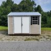 Remove any quotation marks from the generated alt text: The 10x12 Ridgeline Shed #051938626, featuring a small beige design with a metal roof, white double doors, and a window adorned with black shutters, is situated in a grassy area surrounded by trees in the background.