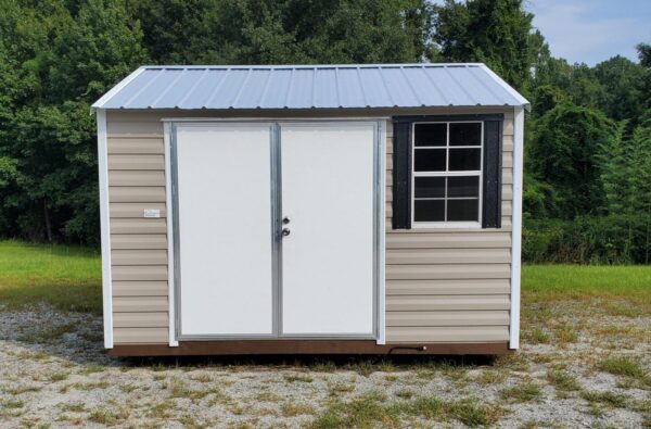 Remove any quotation marks from the generated alt text: The 10x12 Ridgeline Shed #051938626, featuring a small beige design with a metal roof, white double doors, and a window adorned with black shutters, is situated in a grassy area surrounded by trees in the background.