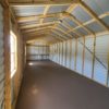 Interior of an unoccupied 12x32 Ridgeline Garage, featuring a long and rectangular layout with high ceilings. The walls and roof are constructed from metal sheets, complemented by visible wooden framing. Sunlight pours through a small window on the left side, projecting a bright rectangle on the floor.