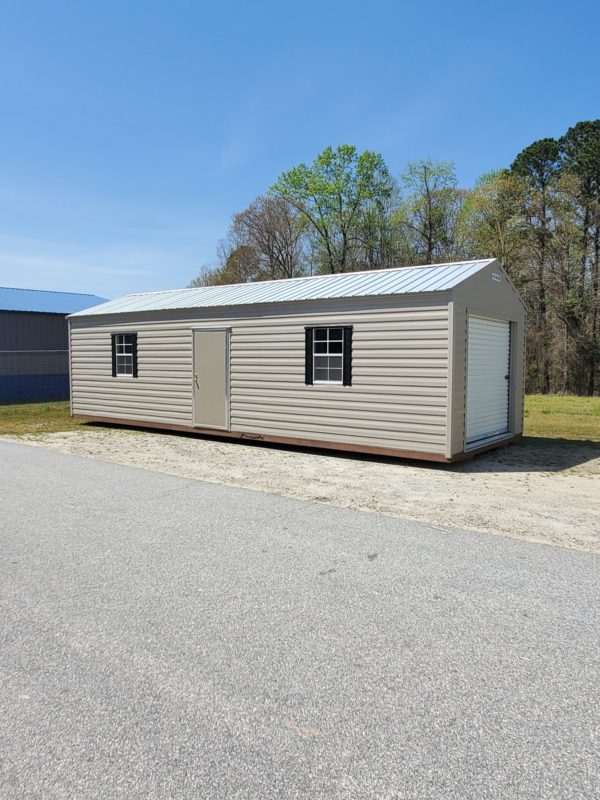 A 12x32 Ridgeline Garage with a beige exterior and metal roof stands on a gravel surface. It features a door and two windows adorned with black shutters, set against a backdrop of trees and a clear blue sky.