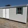 A 12x24 Ridgeline Shed with double white doors, gray roof, and two green-trimmed windows sits on gravel and grass under a blue sky.