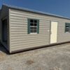 A 12x28 Ridgeline Garage with beige siding, white door, green-trimmed windows, open entry on gravel, grass and trees nearby.