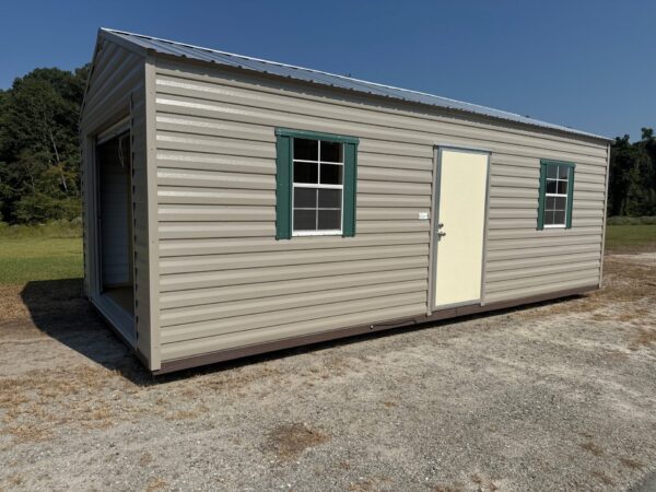 A 12x28 Ridgeline Garage with beige siding, white door, green-trimmed windows, open entry on gravel, grass and trees nearby.