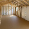 Interior of an empty 12x24 Ridgeline Shed with exposed wood framing, metal roof, one window, and a closed white door. Sunlight streams in.