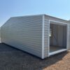 A 12x20 storage garage shed with white trim and light gray metal exterior stands open on dry grass, with trees and buildings behind.