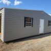 12x20 storage garage shed with light gray metal exterior, white door, two black-trimmed windows, open bay on dry grass under clouds.