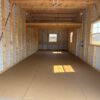 Interior of an empty, unfinished 12x20 storage garage shed with exposed beams, metal wall panels, right-side windows, and sunlight patches on the floor.