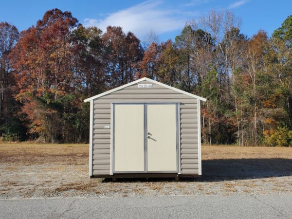 A 10x12 Ridgeline Shed (#083038993) in beige and brown, featuring double doors, is positioned on a gravel area amidst the backdrop of autumn trees. The clear sky is dotted with a few clouds, and the grassy surroundings are adorned with red and orange foliage.