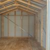 A glimpse inside the 10x12 Ridgeline Shed #083038993 reveals its unfinished wooden walls, plywood floor, exposed beams, and metal braces providing support. Sunlight streams in through an open door, illuminating the pitched roof and enhancing the shed's spacious atmosphere.