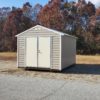 A 10x12 Ridgeline Shed #083038993 in tan and beige with a metal door is situated on a grassy area next to a paved road. Behind the shed, trees display autumn-colored leaves in shades of red, orange, and brown against the backdrop of a blue sky.