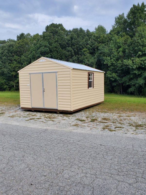 The 12x16 Ridgeline Shed #041138463, featuring a cream color and compact size, sits on a gravel surface. It boasts a gable roof, metal doors, and a solitary window on one side. This shed is nestled against a backdrop of dense green trees under an overcast sky.