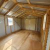 Interior view of the 12x16 Ridgeline Shed #041138463, featuring an empty wooden interior with a corrugated metal roof and plywood floor. Sunlight pours in through windows on both the left and right sides, highlighting the spacious and unfinished area. Large closed doors are visible at the far end.