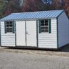 A 12x16 Ridgeline Shed (#083038998) with a small white structure and a blue metal roof features two windows adorned with green shutters and a double door. It is situated on a cleared area beside a roadway, with trees displaying autumn-colored leaves visible in the background.