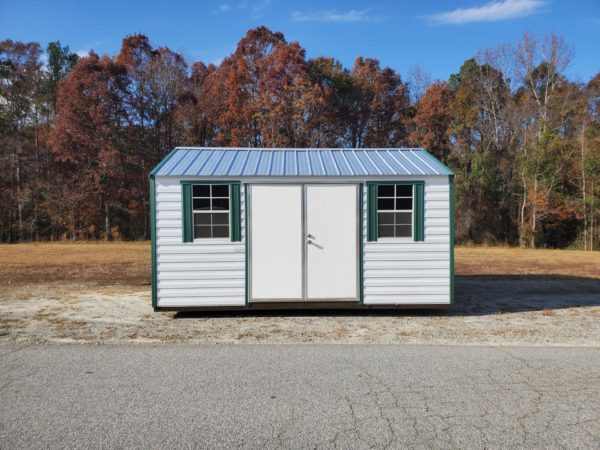 The 12x16 Ridgeline Shed #083038998, featuring a compact design in light green and white with a metal roof, stands in a clearing. It includes two windows adorned with green shutters and double doors. In the background, trees with autumn foliage can be seen under a blue sky dotted with wispy clouds.