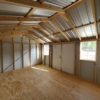 Interior of an unoccupied 12x16 Ridgeline Shed #083038998 with unfinished wooden walls and a metal roof. Sunlight streams in through two windows, illuminating the plywood floor. A door provides access to the exterior. The space is bright and roomy.