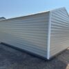 A 12x16 beige Ridgeline Shed (#041138463) with a sloped roof stands on dry grass, another similar shed behind it under blue sky.
