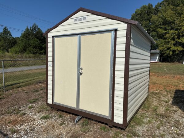 An 8x10 beige and brown Ridgeline Shed with double doors stands on gravel in a grassy yard bordered by a fence and trees.