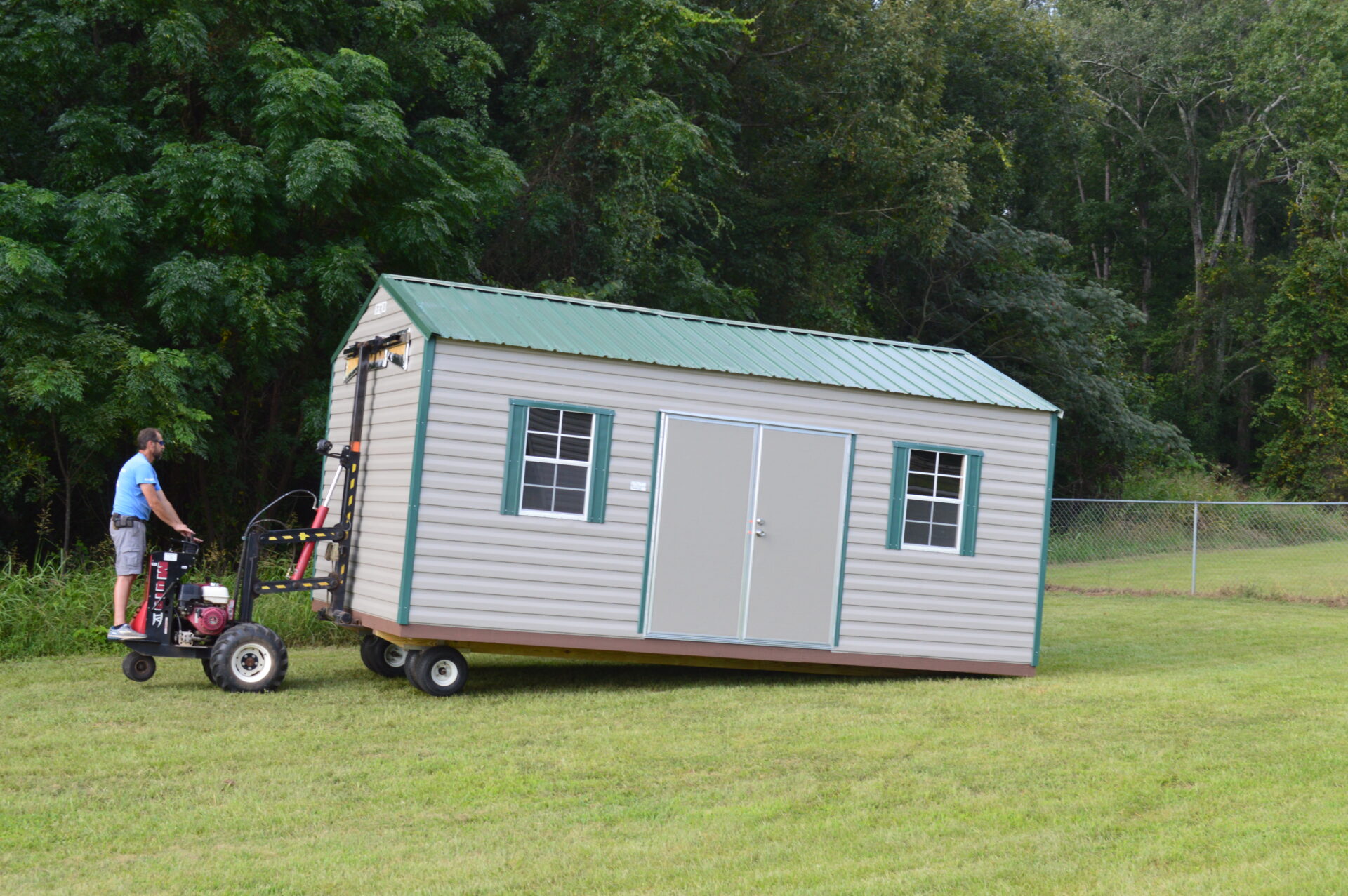 A person maneuvers a small gray and green shed on wheels using a motorized dolly across a grassy area, with trees and a chain-link fence in the background.