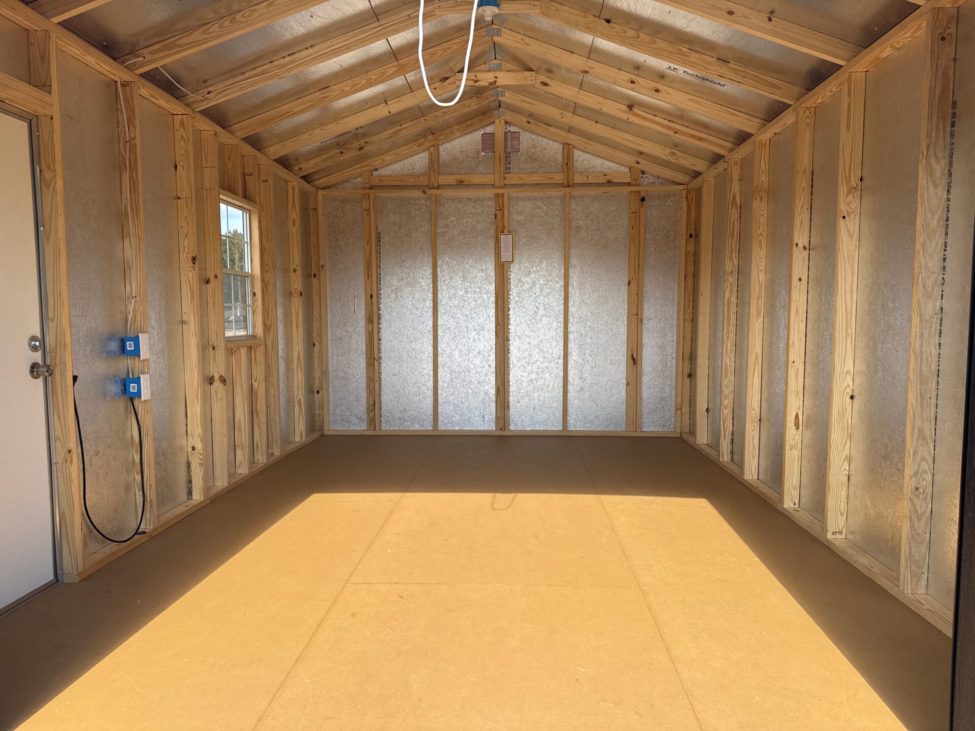 Interior of empty 10x16 Frontier Shed with exposed beams, insulation, closed white door, blue outlets, and sunlight on floor.