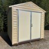 8x8 Ridgeline Shed, beige metal with double doors and gable roof, sits on gravel among grass and bamboo. Available now.