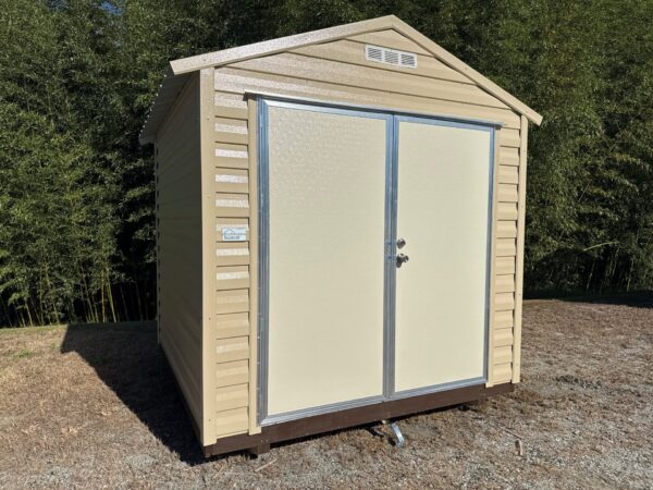 8x8 Ridgeline Shed, beige metal with double doors and gable roof, sits on gravel among grass and bamboo. Available now.